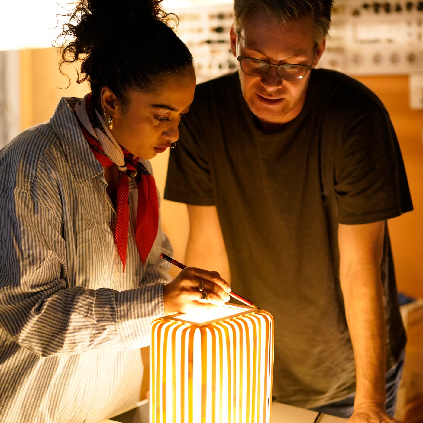 Two people reviewing a glowing bamboo lamp prototype, adjusting details and examining the light in a workshop setting.