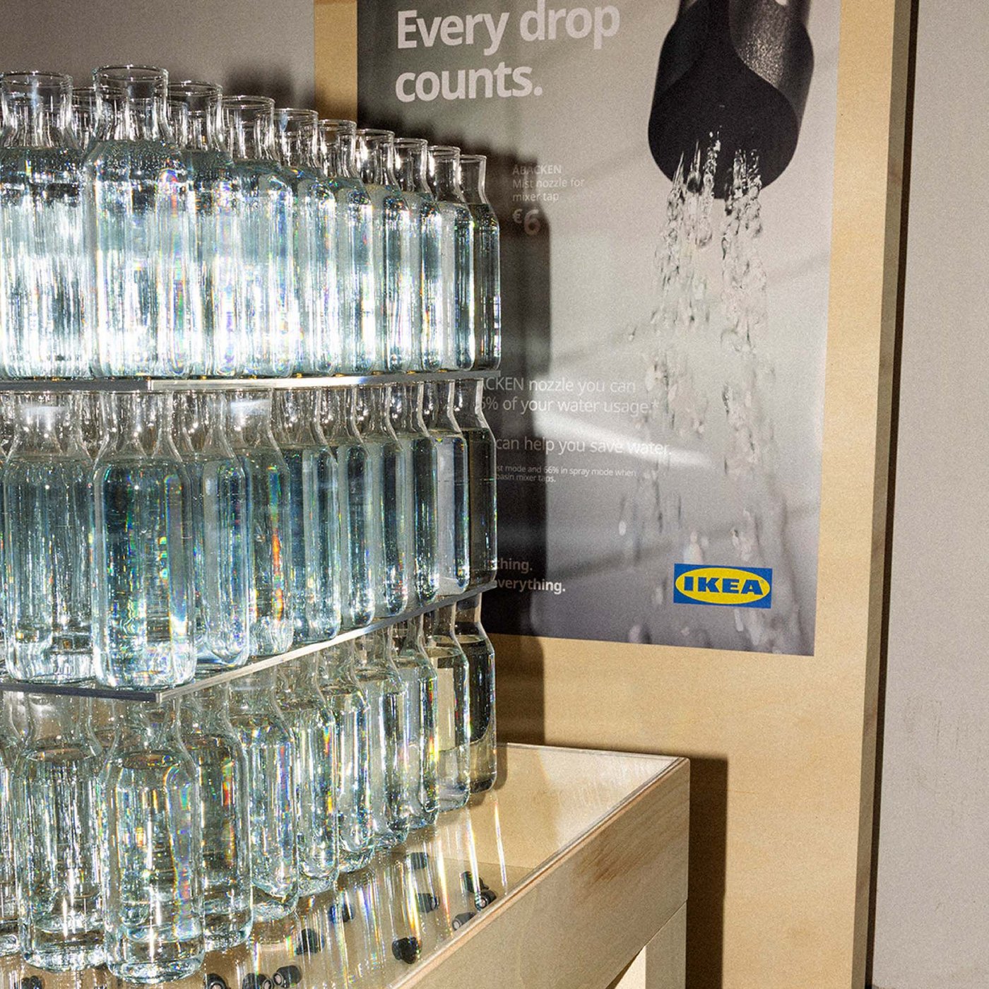 Lots of clear bottles filled with water, arranged in three stacks in front of an IKEA poster.