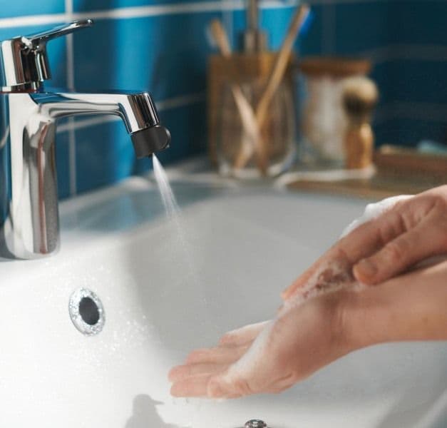 A bathroom sink and two soapy hands being sprayed with just a little water by an ÅBÄCKEN mist nozzle for mixer tap.