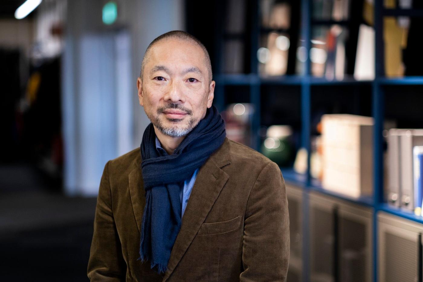 Yasushi Kusume looks at the camera. behind him is a bookshelf along the wall, filled with objects.