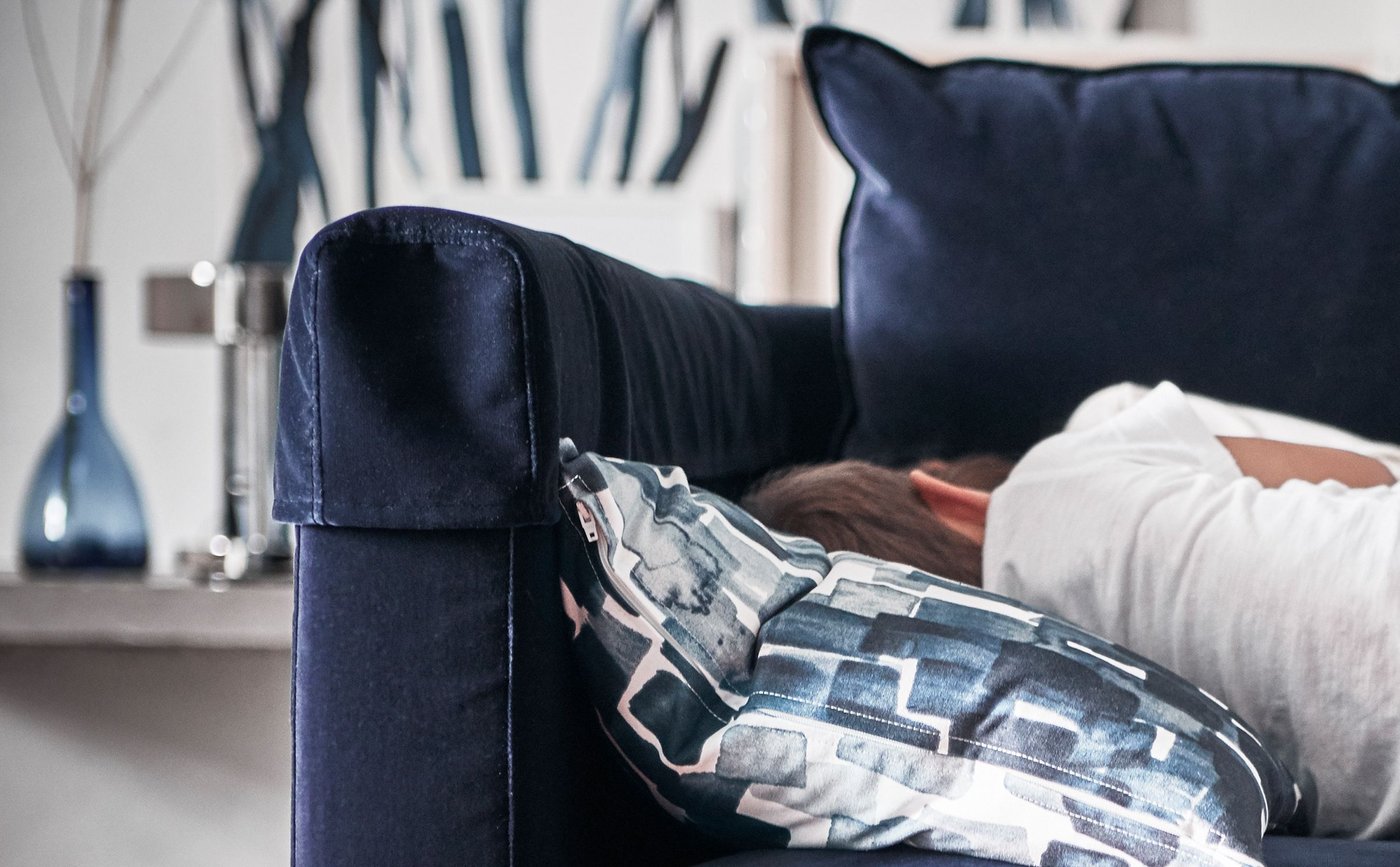 A child in jeans and a white t-shirt sleeps on a blue sofa, with his head on a blue and white patterned cushion.