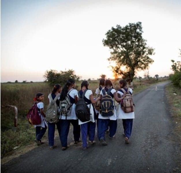 A group of girls carrying backpacks are walking on a dirt road. Their hands are placed on each other’s backs.