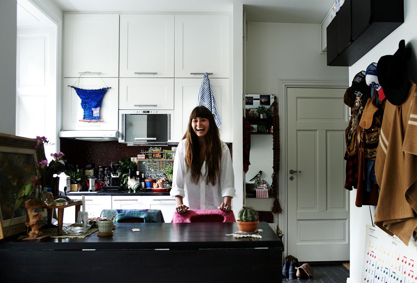 A woman with long brown hair laughs as she stands behind a black kitchen island. Behind her is a white cosy kitchen.