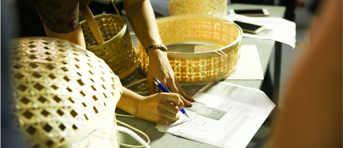 A person is leaning over a table to make notes on a piece of paper. They are surrounded by woven lampshades of various sizes.
