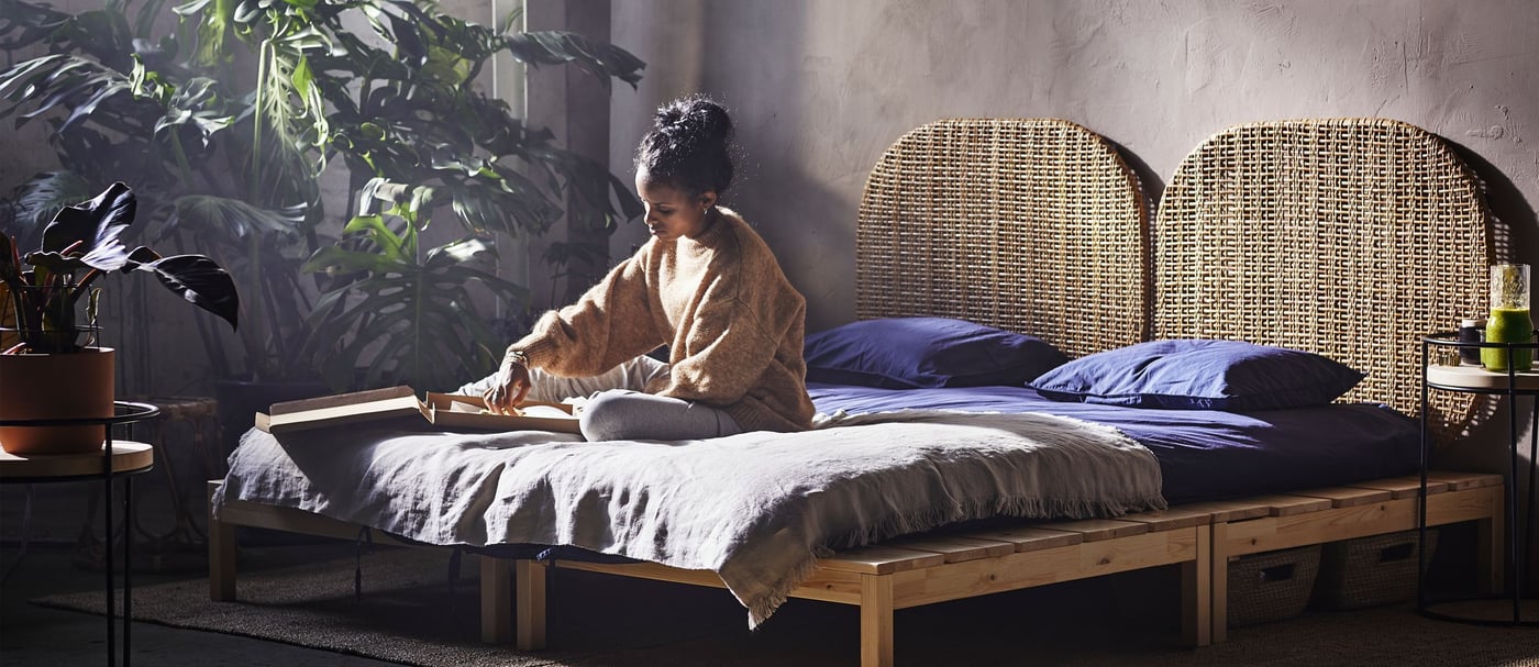 A woman sitting on a bed with a rattan headboard, surrounded by lush plants.