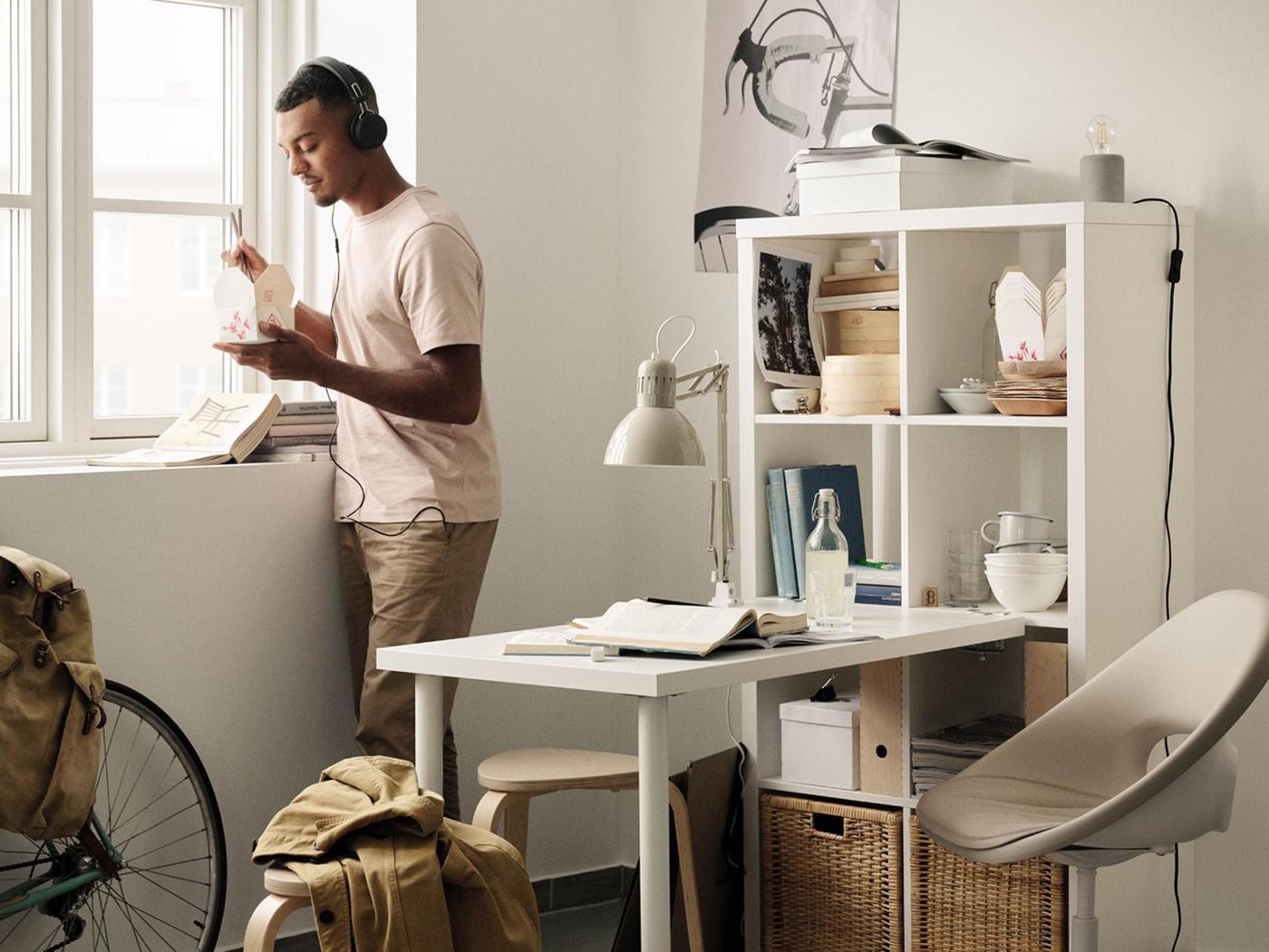 A young man wearing headphones is standing by the window in his home, eating food. In his room is a bicycle and a desk.