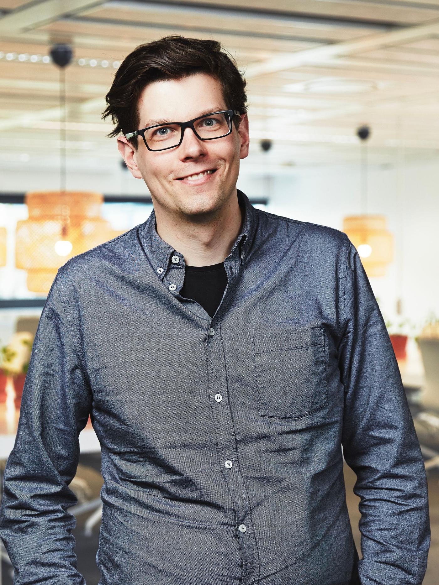 A man in a blue shirt and black glasses standing in an office environment.