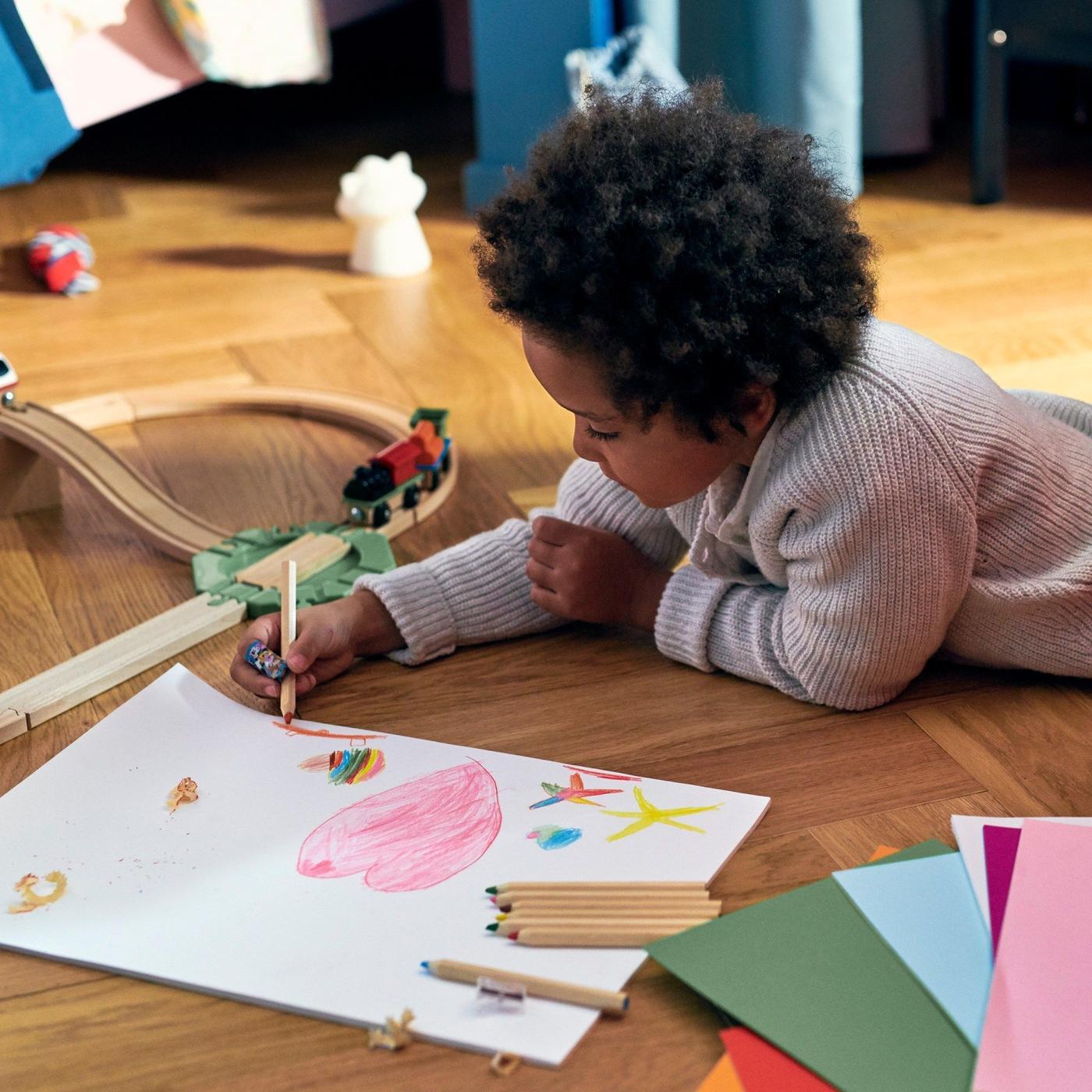 A young child draws a picture while lying on a wooden parquetry floor next to a toy train set.