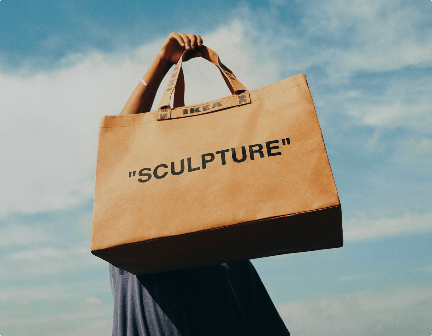 A person holds a brown bag with the word "sculpture". Their face is obstructed by the bag and behind is a blue sky with clouds.