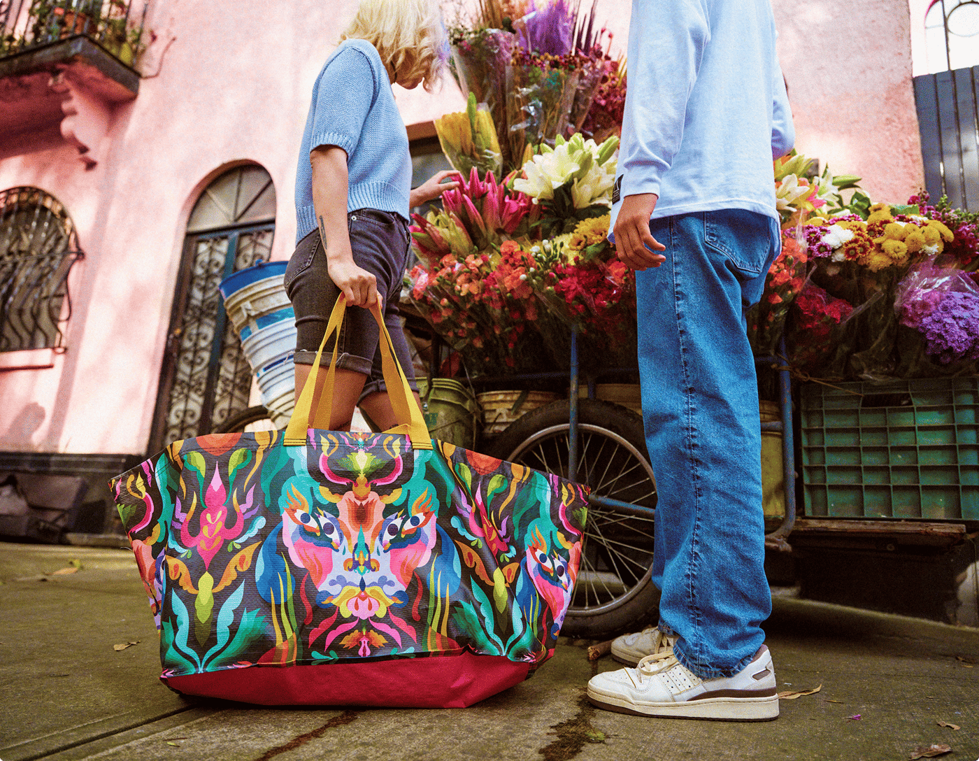 Two people stand outside a pink building with a brightly decorated bag on the ground.