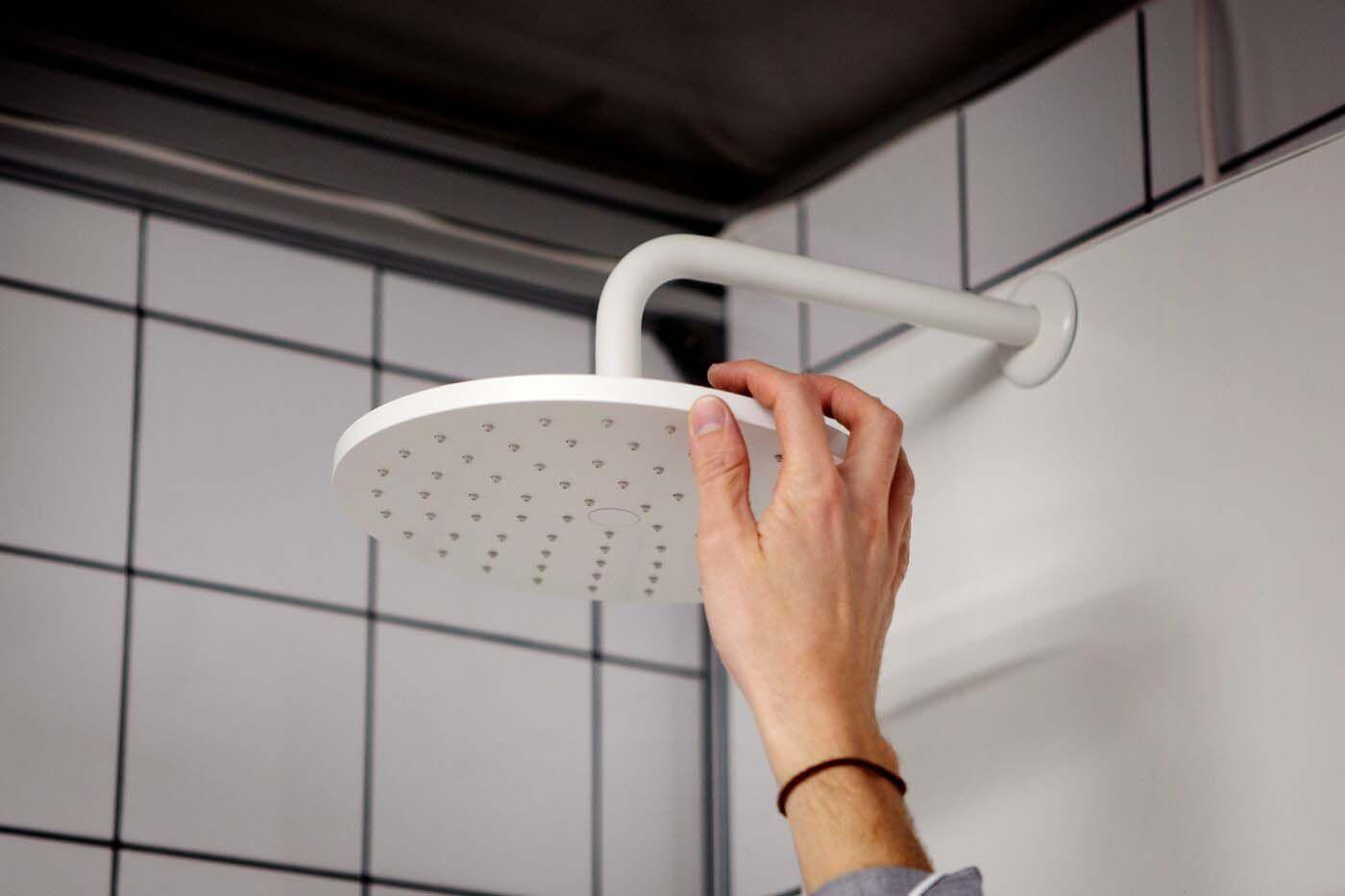 A hand reaches up to touch a white shower head attached to a white wall.