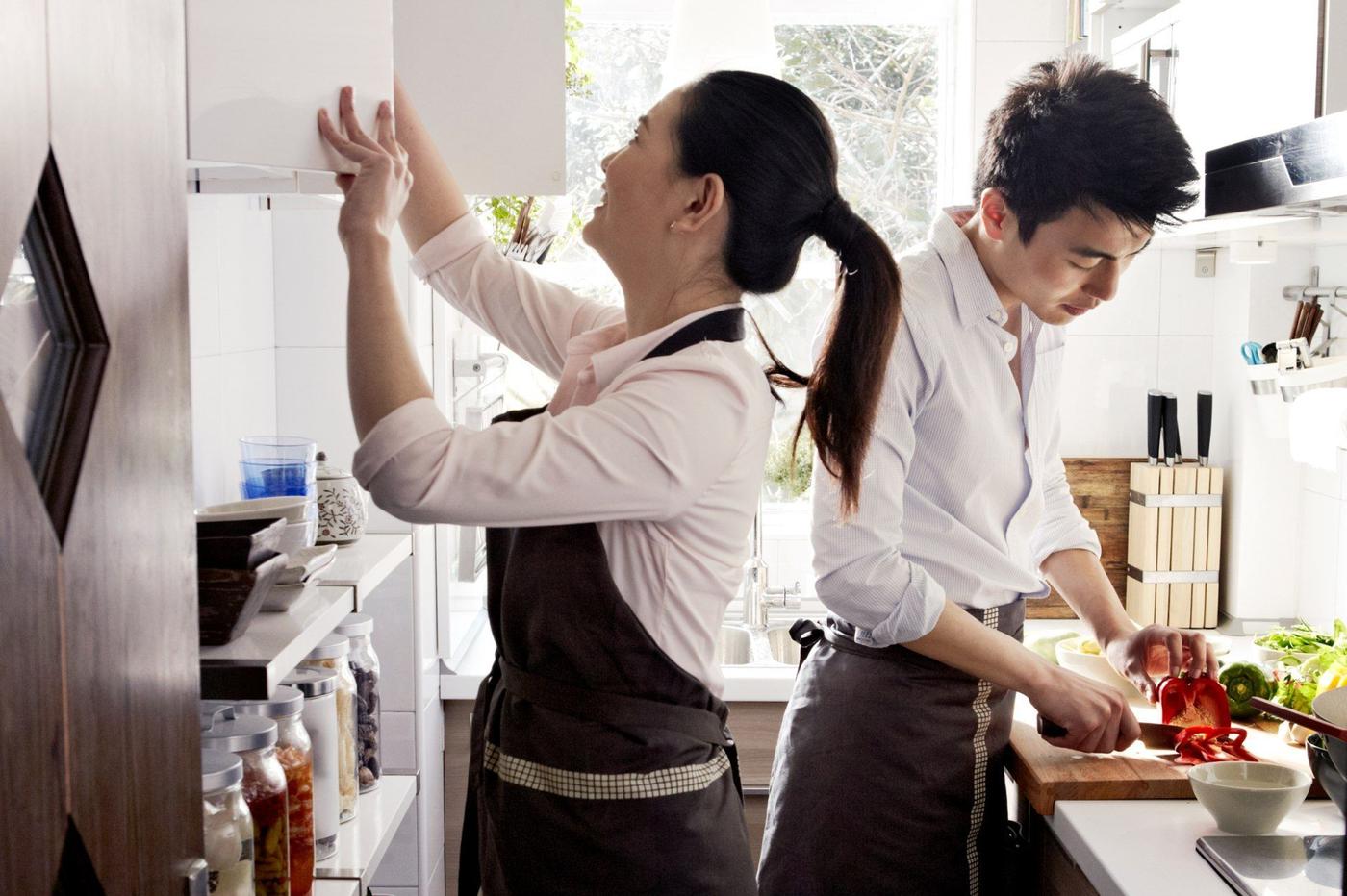 A man and woman cook in the kitchen. The woman is looking in the overhead cupboard.