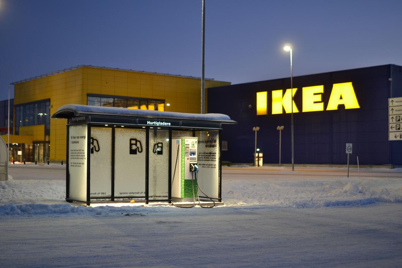An electric charging station lit up by lamps in front of an IKEA store at night. The ground is covered with snow.