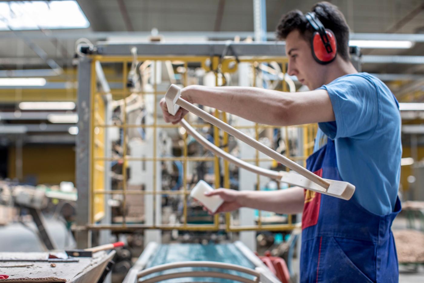 A factory co-worker with blue carpenter’s trousers and a noise cancelling headset is treating a wooden frame.