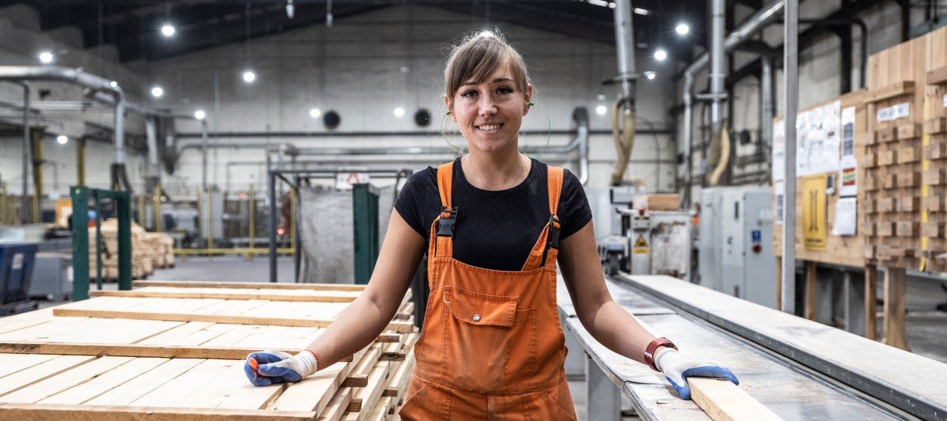 An IKEA co-worker in orange carpenter’s trousers and gloves in a factory, standing next to a large pile of wooden planks.