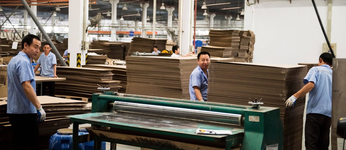 A couple of factory workers wearing light blue shirts and white gloves are working with stacks of cardboard.