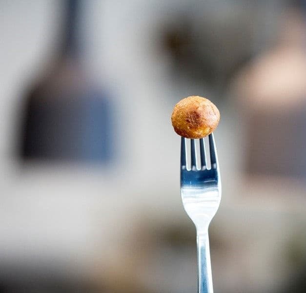 A stainless steel fork stuck into a light brown, fried plant ball and a blurry background.