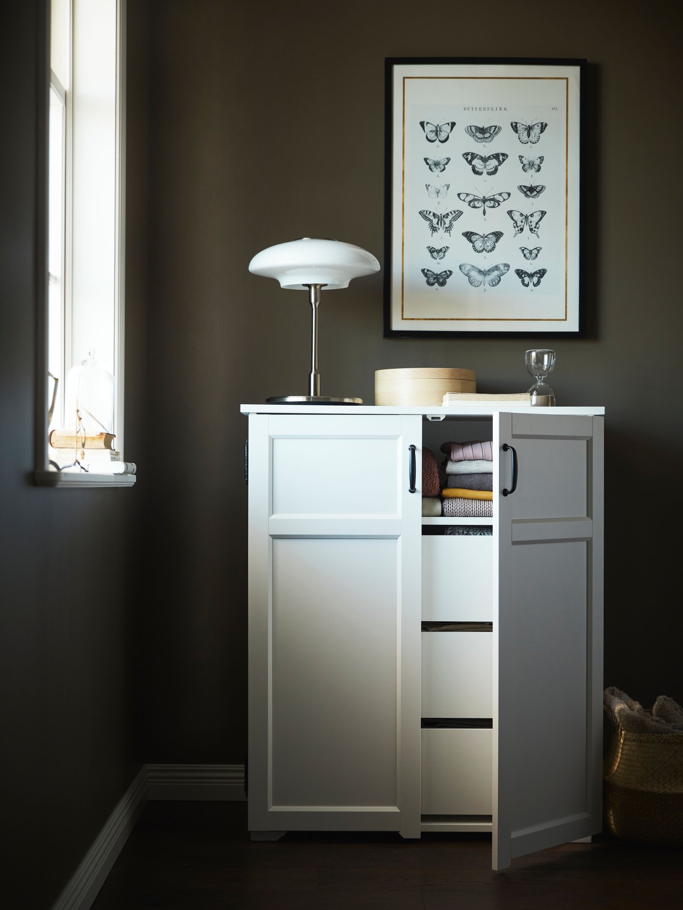 A white cabinet with the door open showing drawers. There is a lamp on top and a grey wall behind.