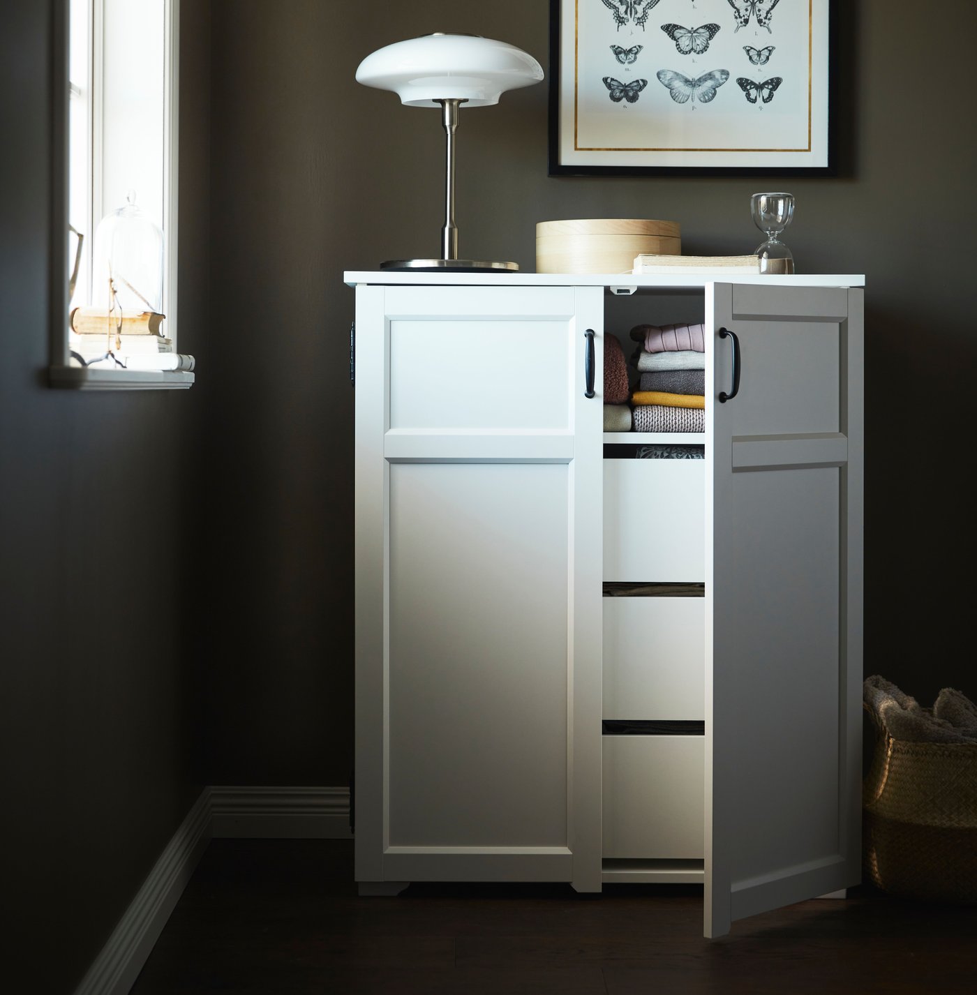 A white cabinet with the door open showing drawers. There is a lamp on top and a grey wall behind.