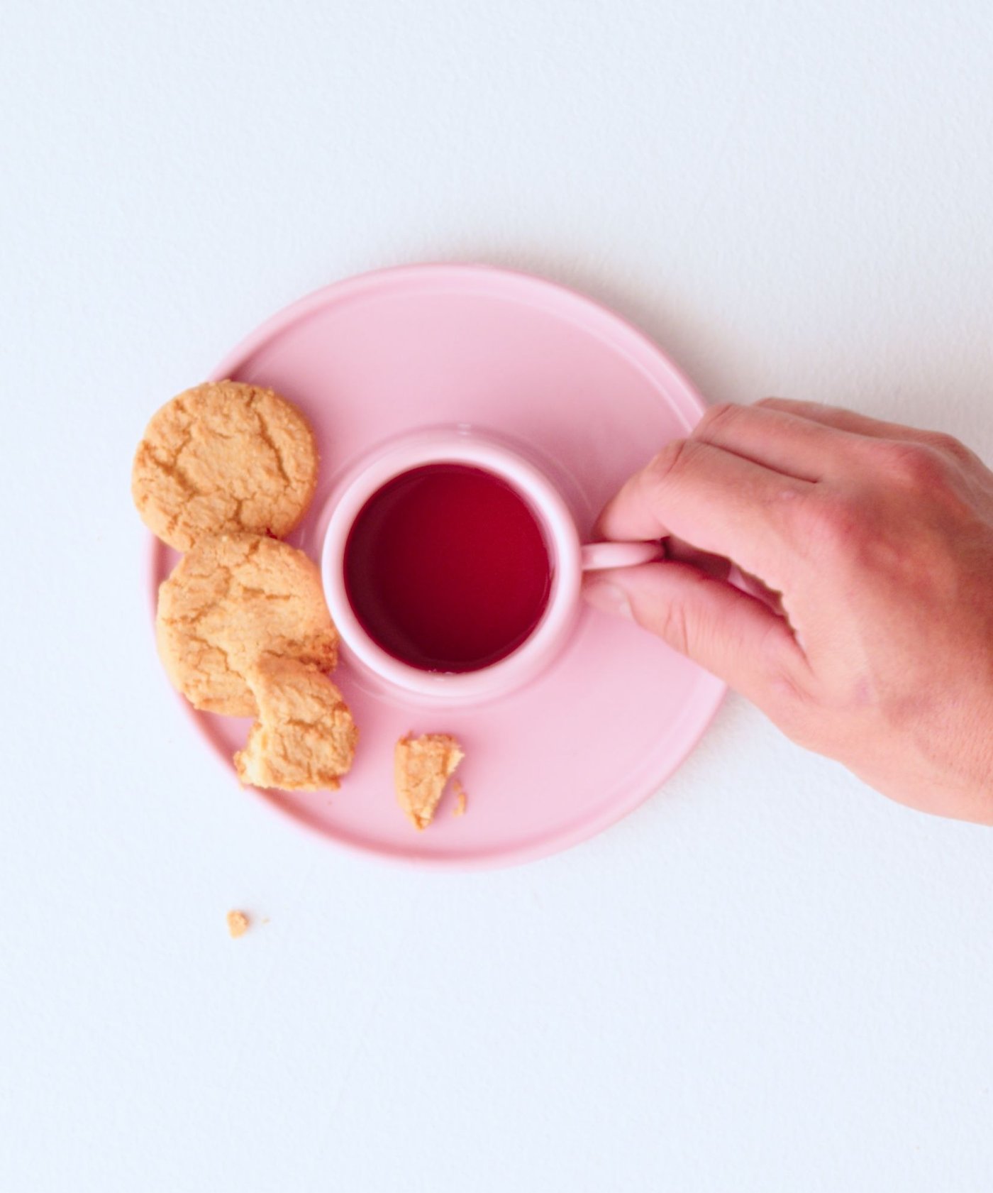 A hand holds the handle of a pink cup holding tea, viewed from a top down angle. On the pink saucer that the teacup is resting on there are three cookies. One has a bite taken from it.