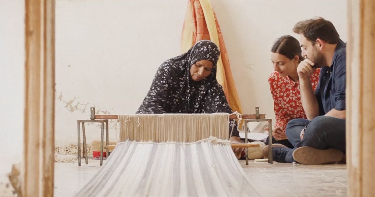 Two people watching a woman weave.