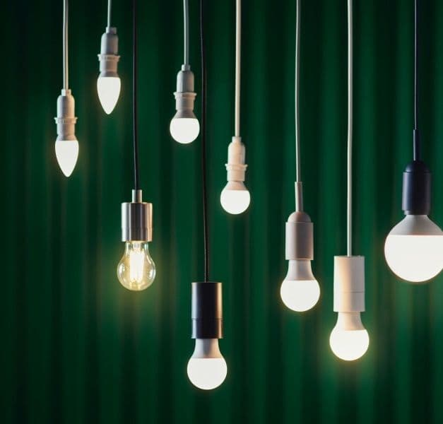 Lit LED light bulbs in different shapes and sizes hanging from the ceiling at different heights in front of a green curtain.
