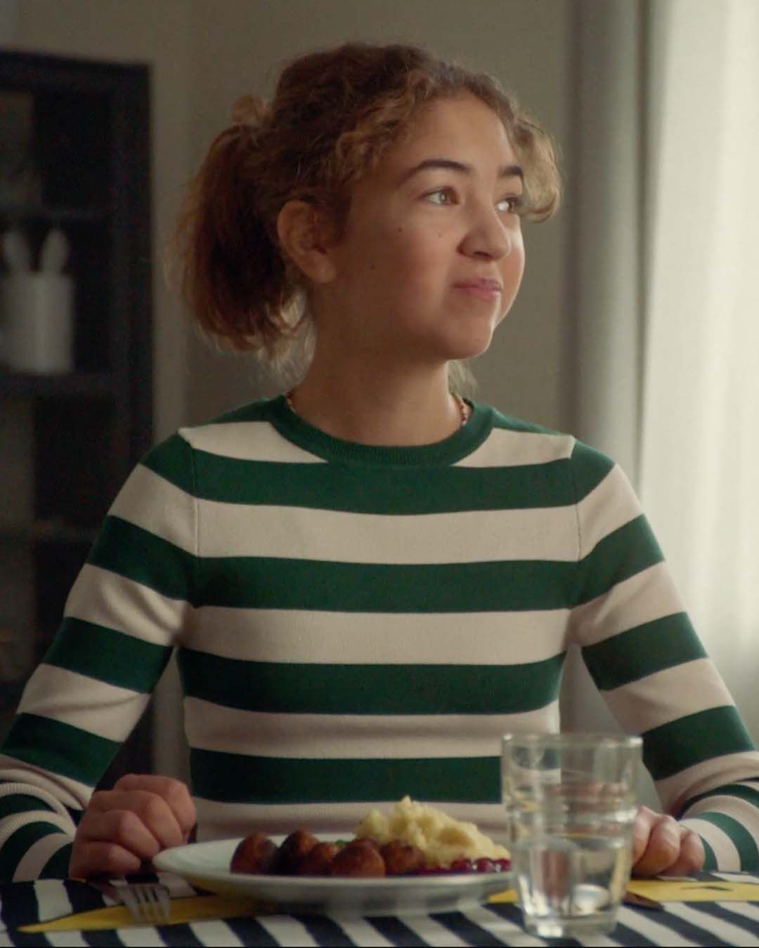 A teenage in a striped top looks thoughtful. She is eating meatballs with peas and mashed potato.