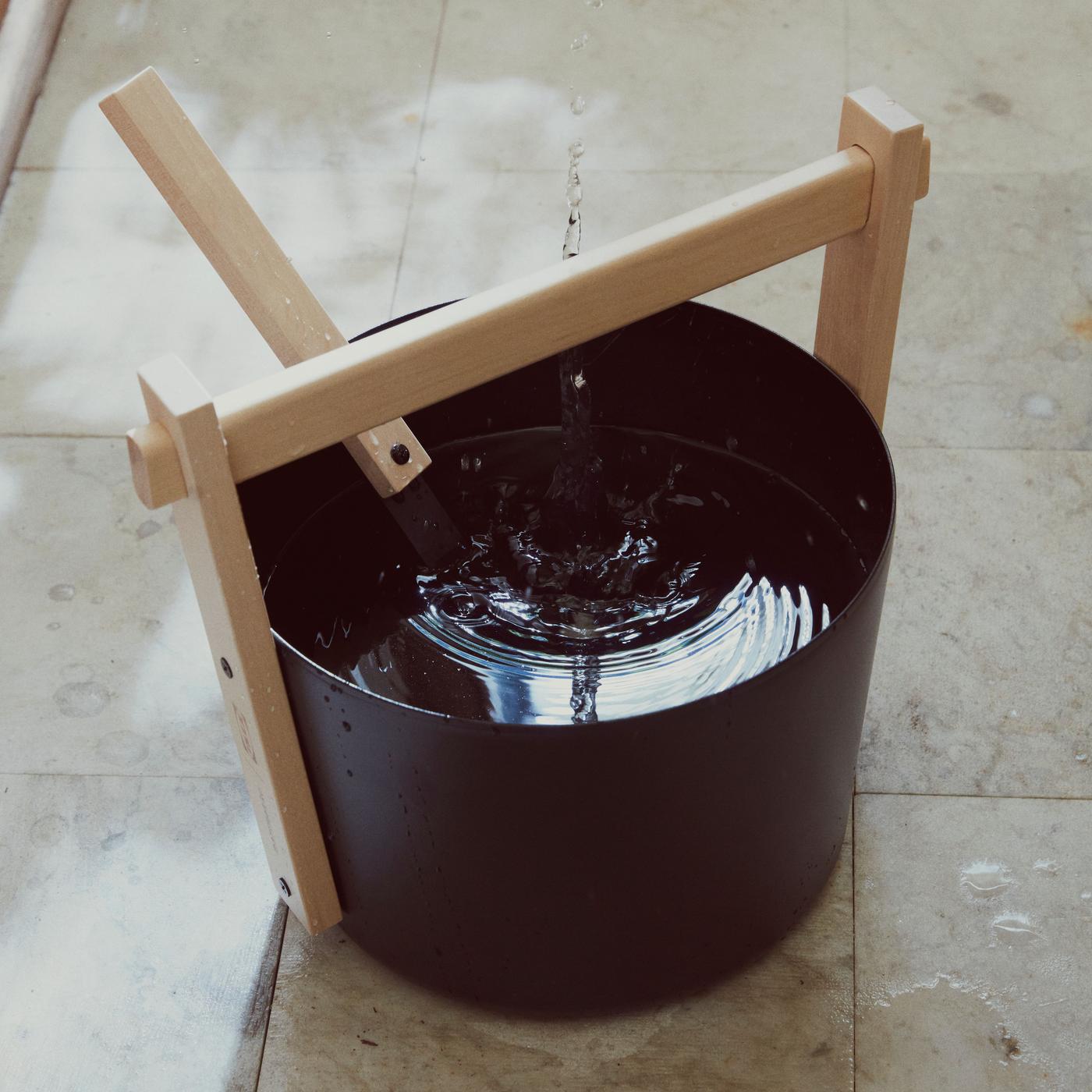 A black sauna bucket with a wooden ladle standing on the floor of a sauna. 