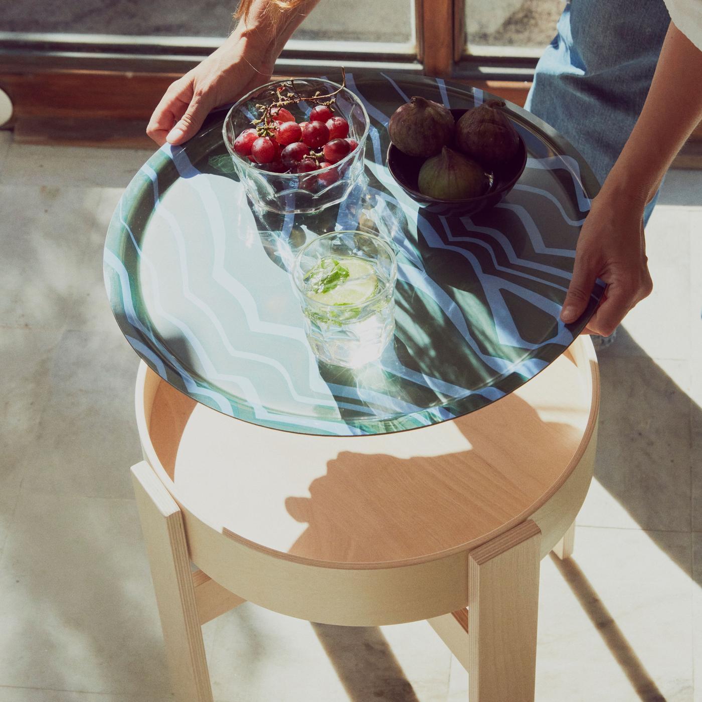 Two hands putting down a tray with grapes, figs and a drink on a side table.