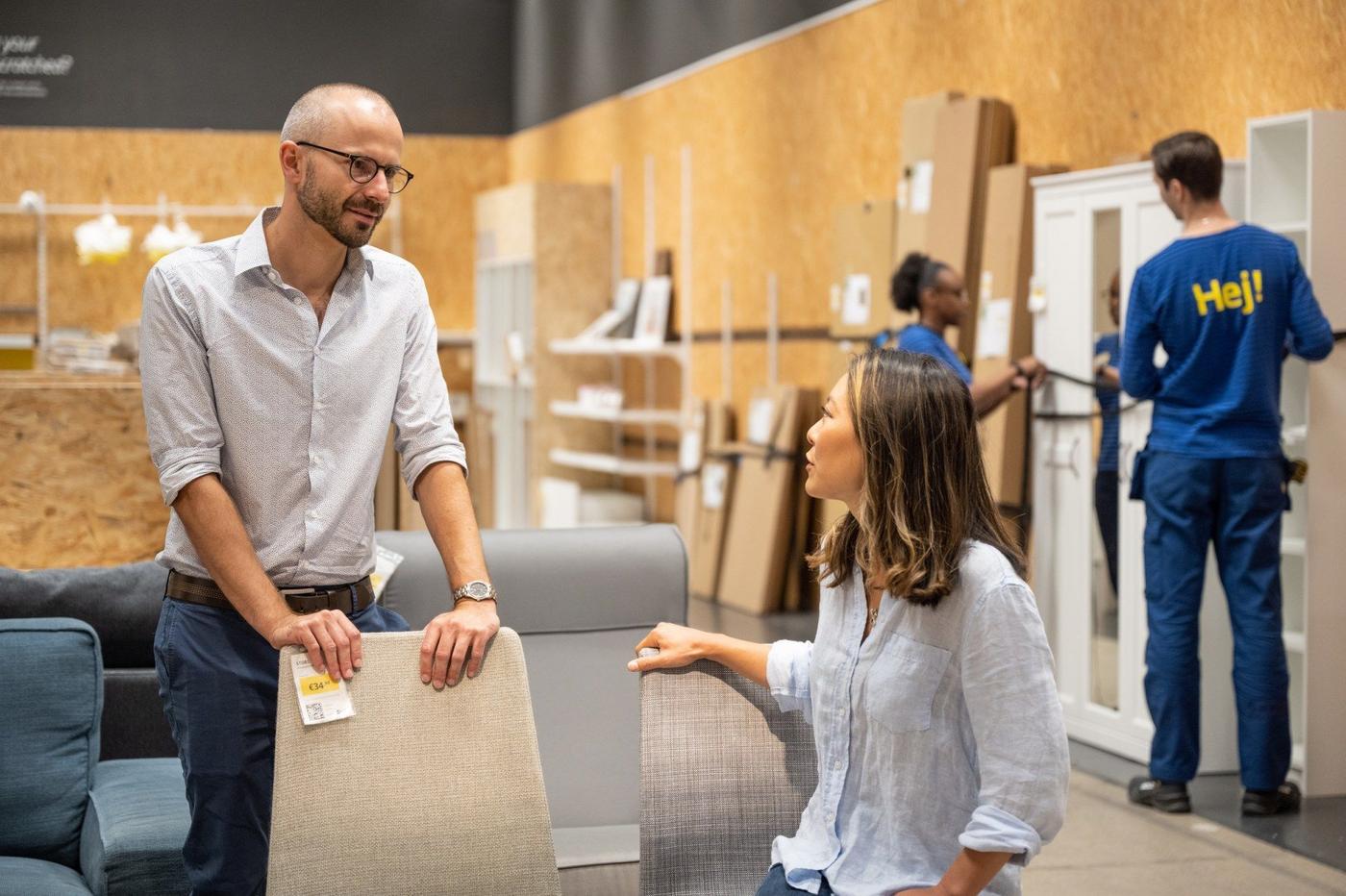 A man stands, holding the back of a chair, while a woman sits and talks to him.