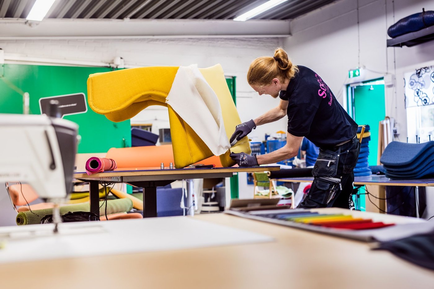 An IKEA co-worker wearing a black t-shirt and pants reupholstering an armchair with bright yellow fabric.