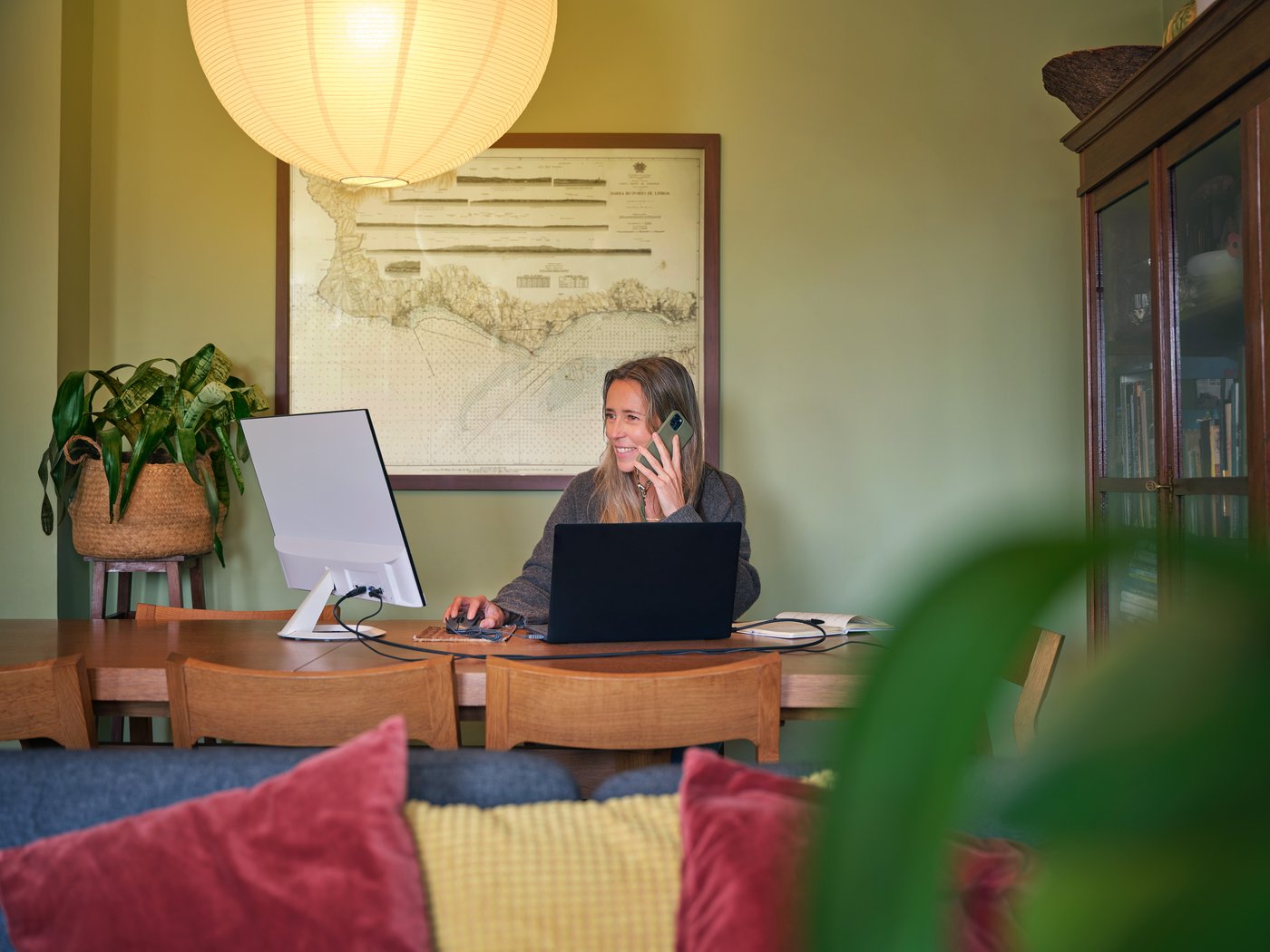 A person sits and works at a computer at a dining table while taking a phone call. 