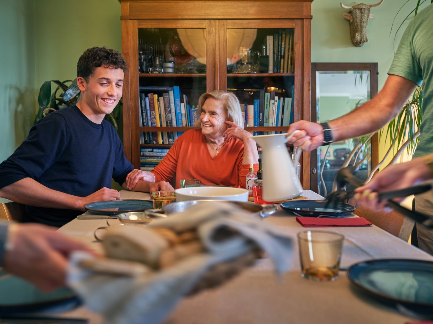A mutli-generational family gather around a dining table, which is being set for a meal.