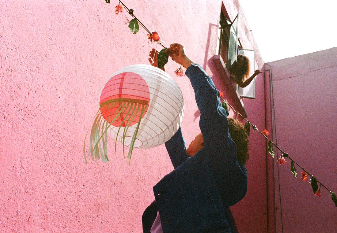 A person hanging a white lampshade on a line in a backyard while another person looks out from a window.
