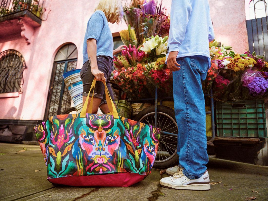Two persons standing at a sidewalk looking at flowers. One of them is holding a colourful bag.