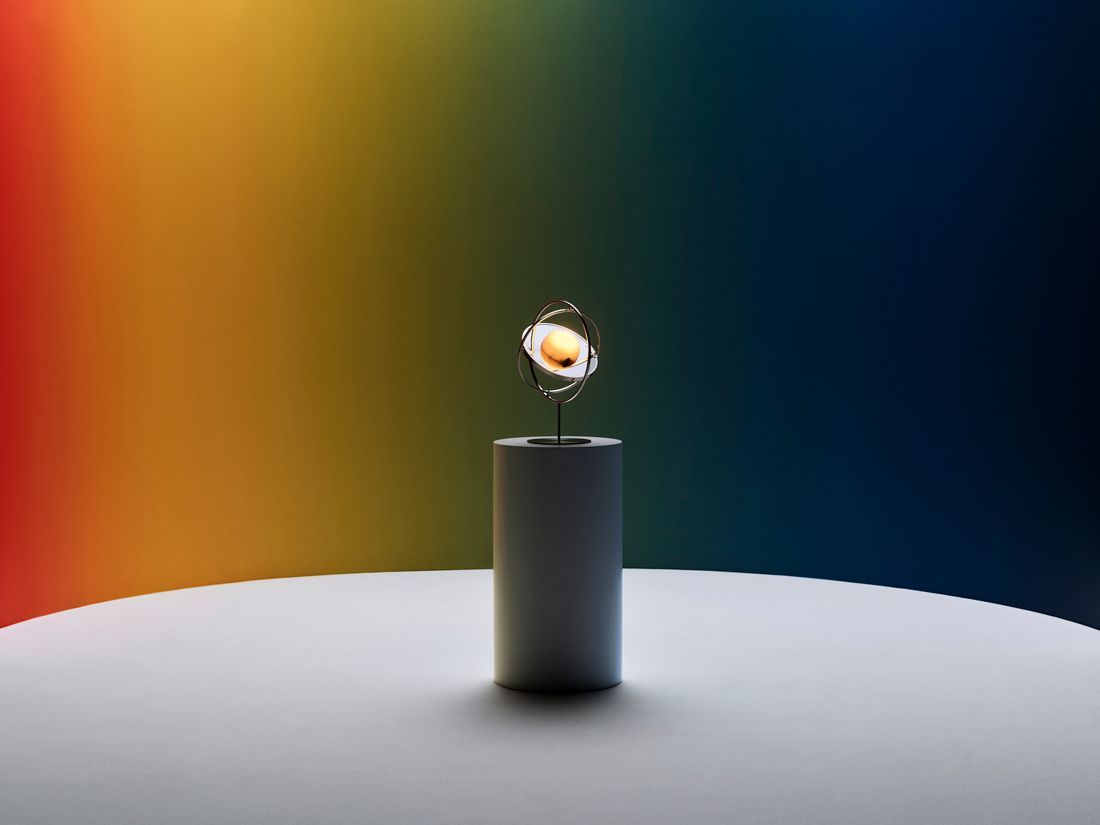 A solar powered stainless lamp with a mirror standing on a podium before a wall painted in the colour of a rainbow.