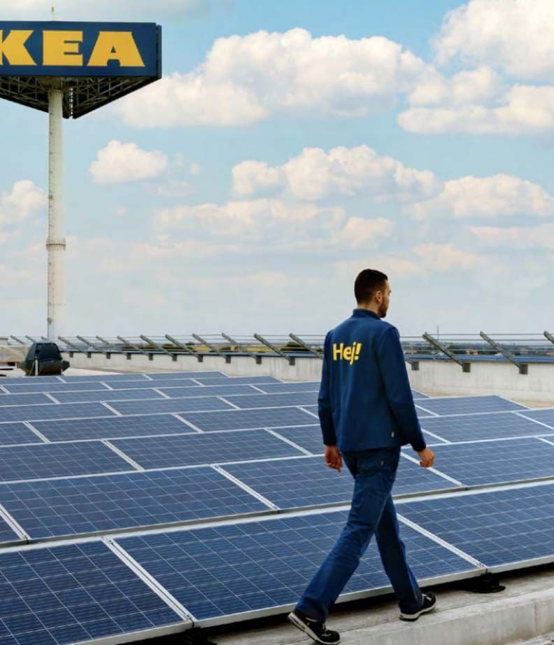 An IKEA co-worker walking outdoors among rows of solar panels, with a large IKEA store sign in the background.