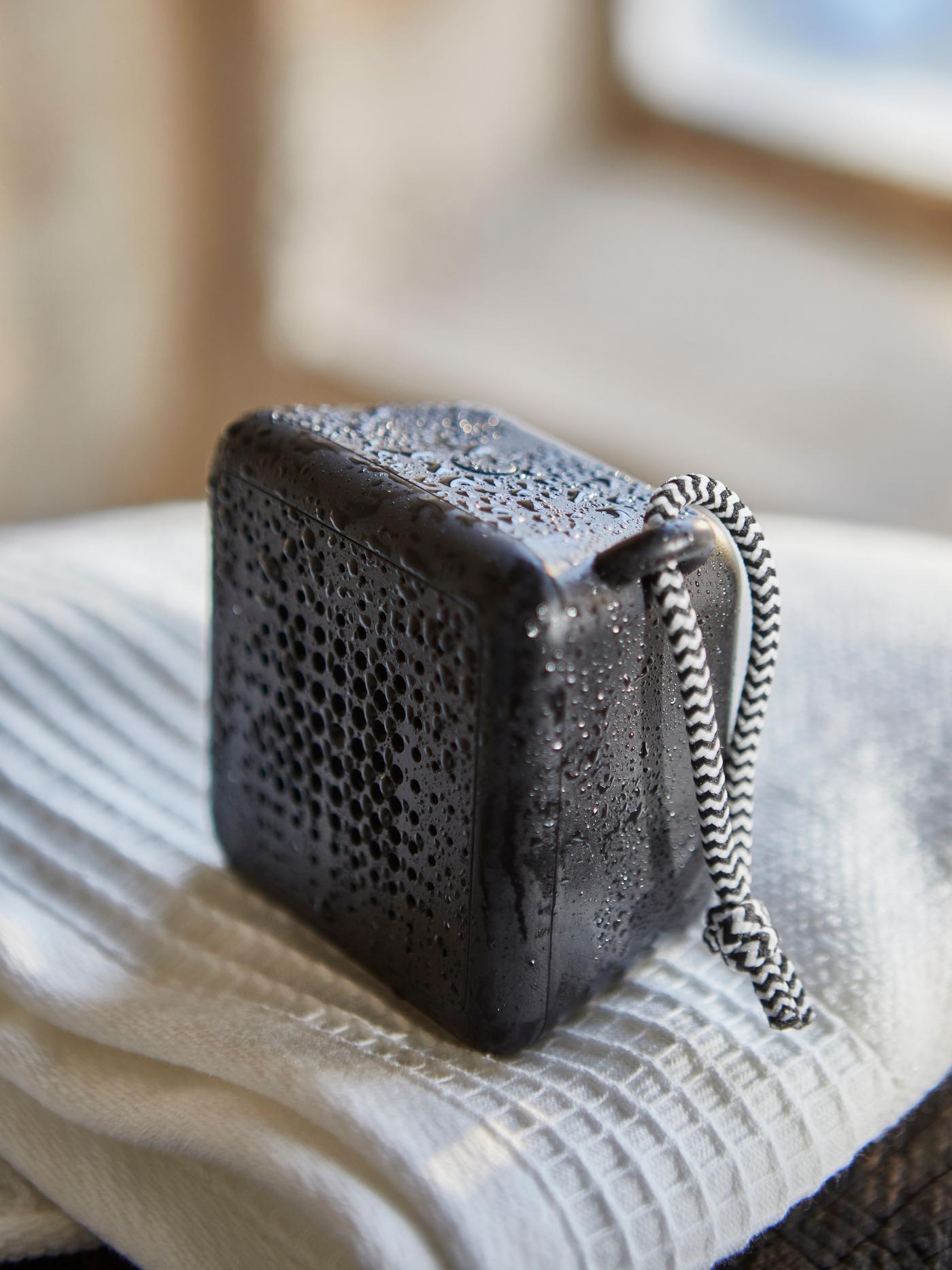 A black waterproof bluetooth speaker, covered with mist, standing on a white towel.