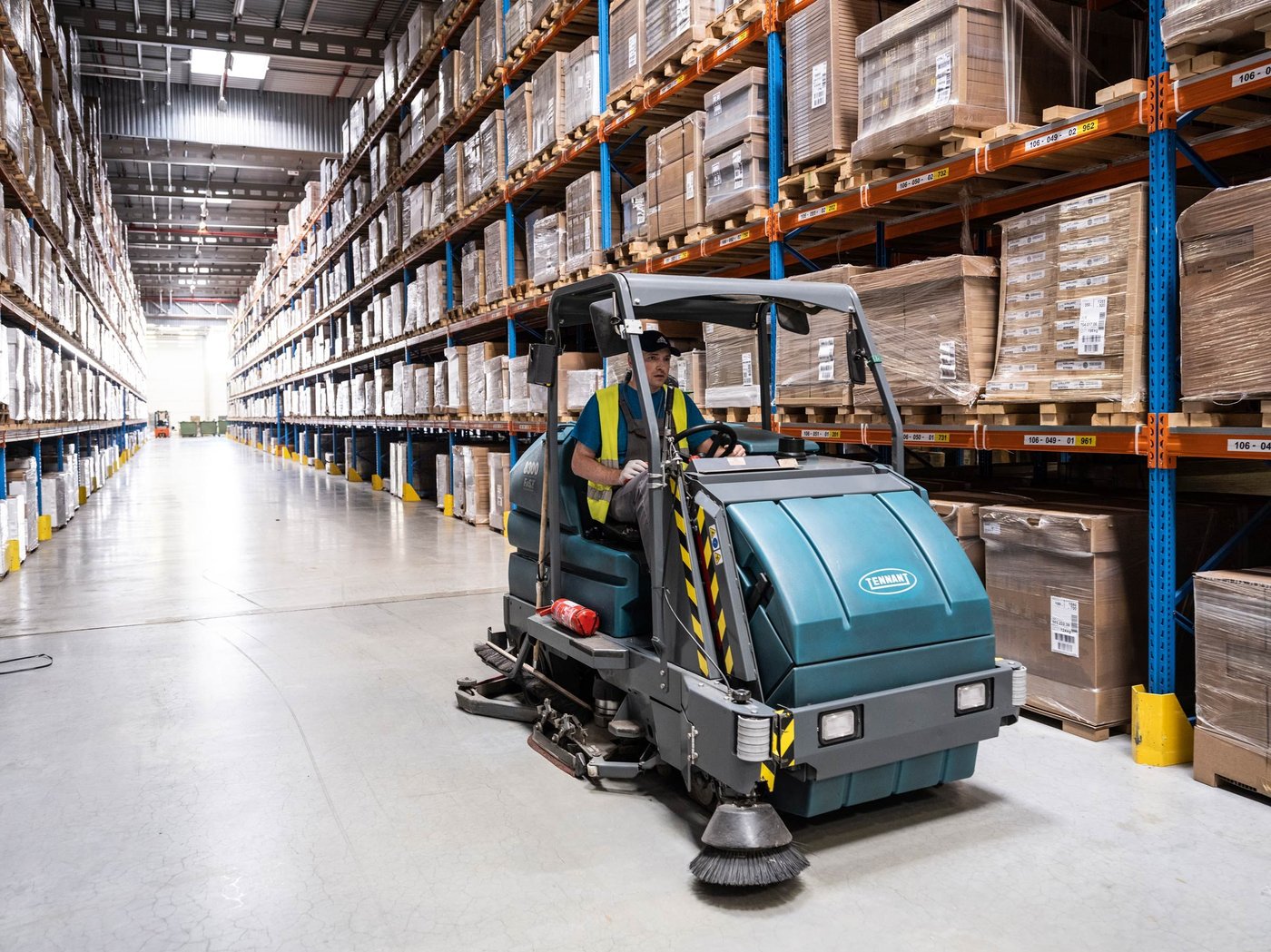 A co-worker in a yellow safety vest drives a cleaning machine through an IKEA warehouse, surrounded by packed IKEA products.