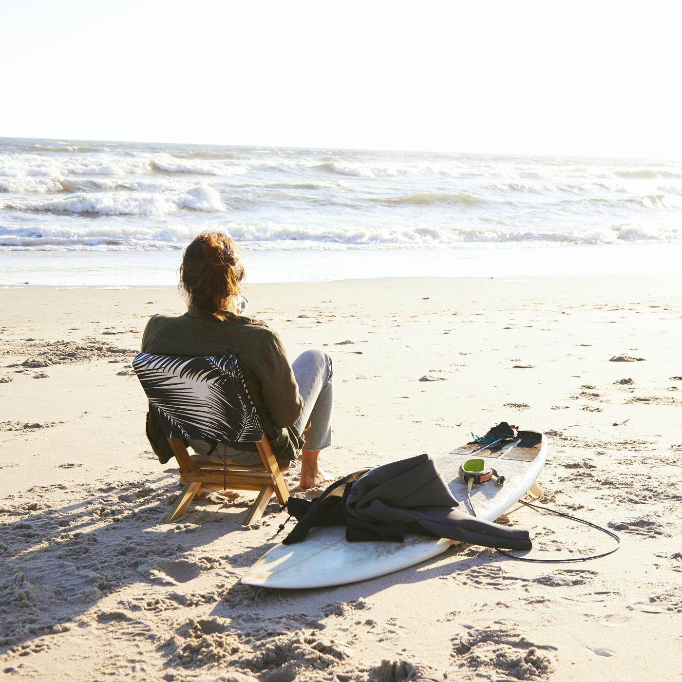 A man sitting in a beach chair, with a surf board next to him, looking at the ocean.