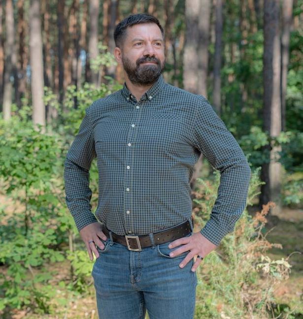 Maciej stands in a forest with his hands on his hips.