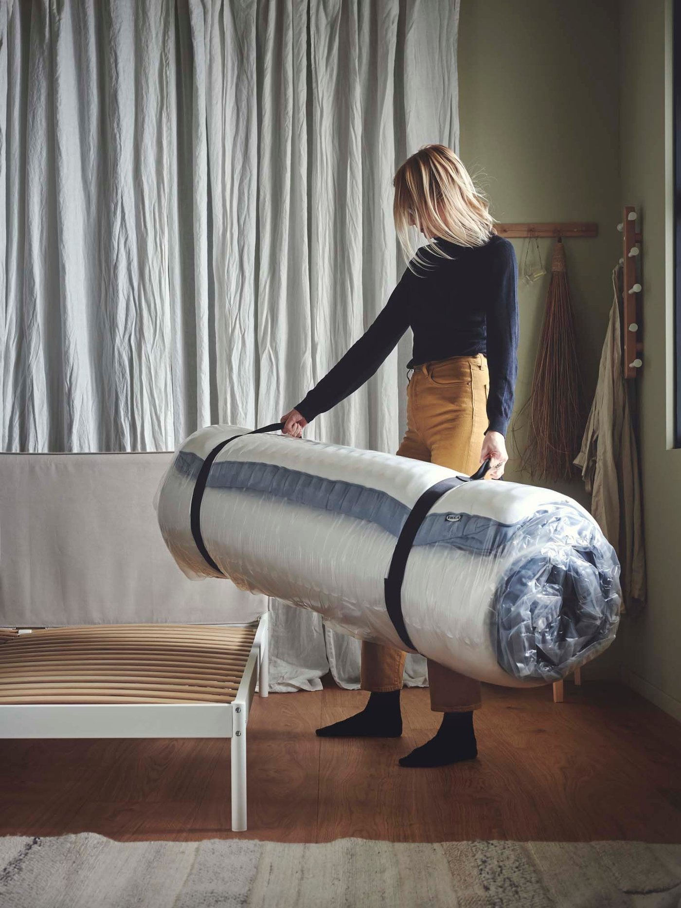 A person stands next to a bed frame, holding a rolled up mattress.