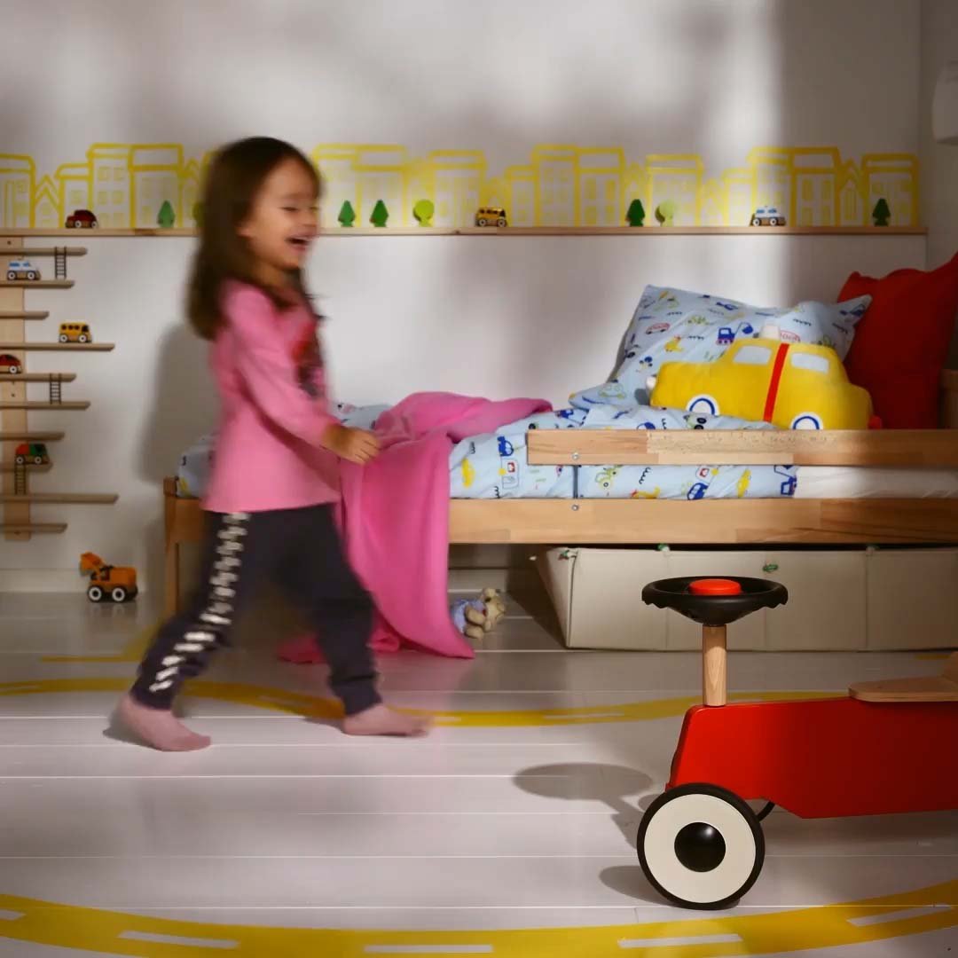 A child in a white and yellow children's bedroom with a red ride-on toy car.