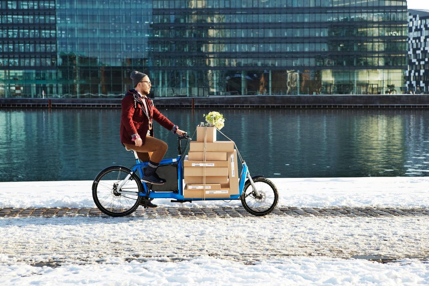 A man transporting IKEA flatpacks on an IKEA rental cargo bike alongside a canal and a glass facade building.