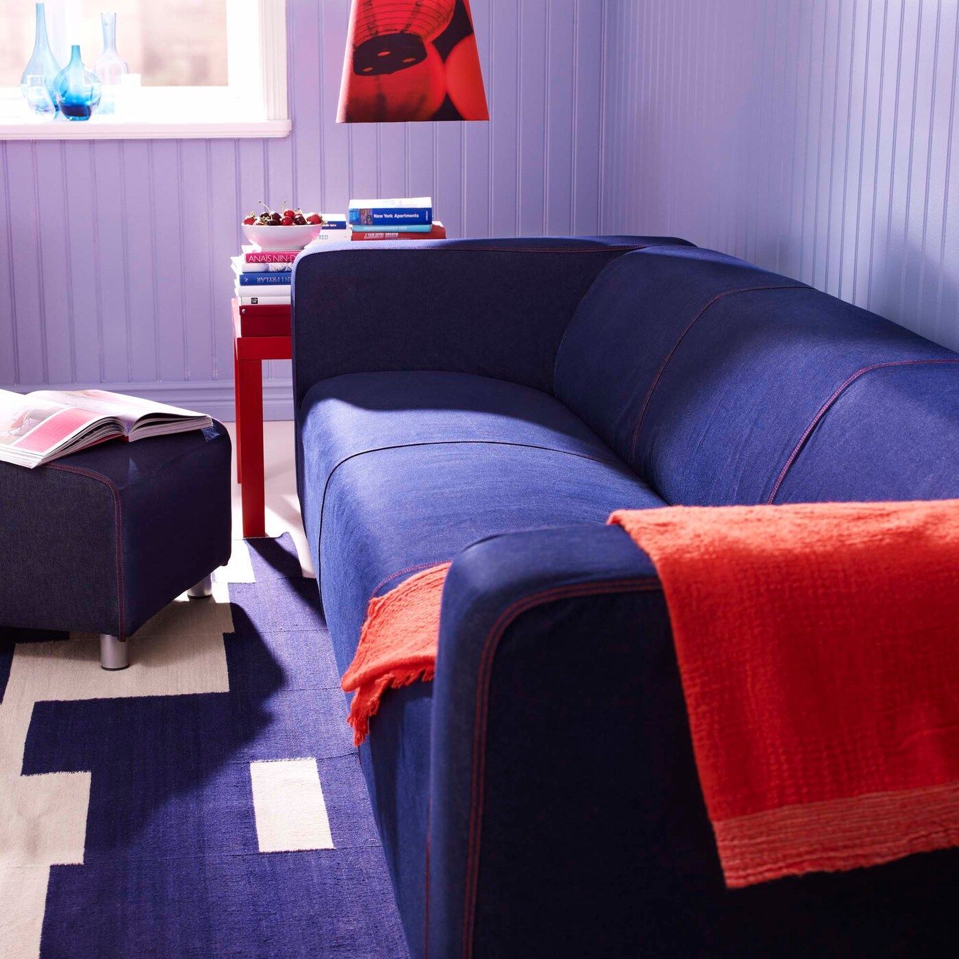 A dark blue denim KLIPPAN sofa in a pale purple room with red throw and side table.