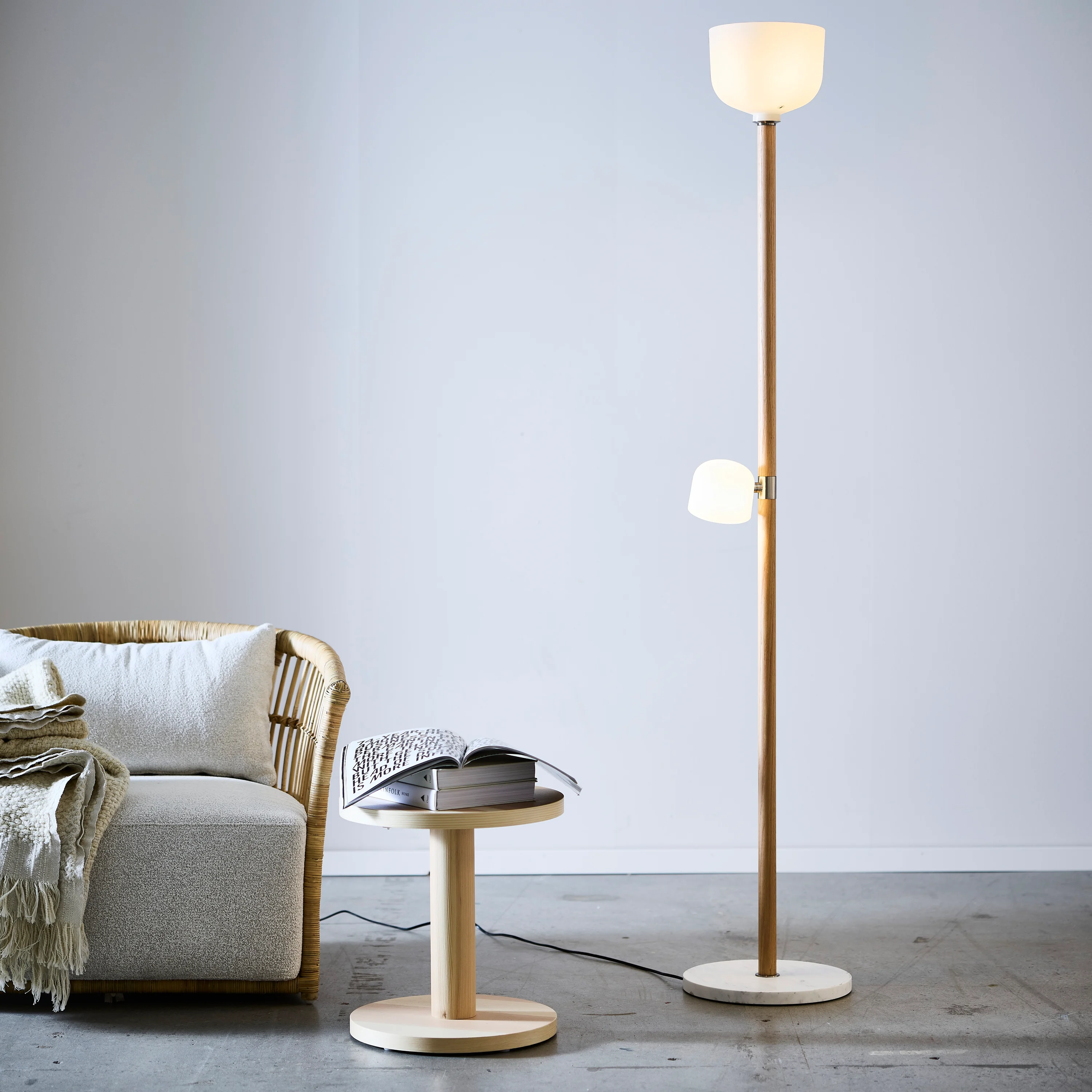 An illuminated floor lamp stands next to a round wood table and an upholstered rattan chair, in a white room with an industrial floor.