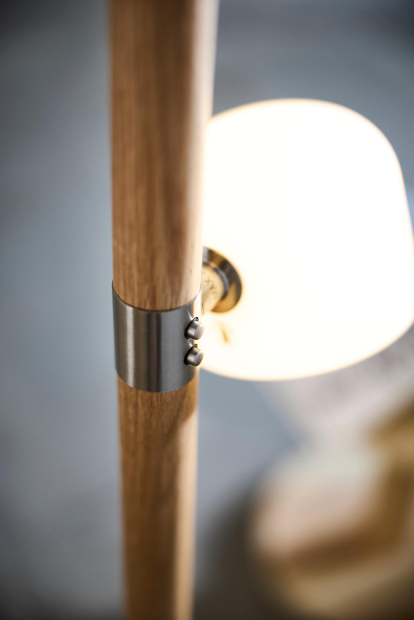 Close-up of wooden lamp stand with metal bracket and buttons, with softly glowing white lamp shade in the background.