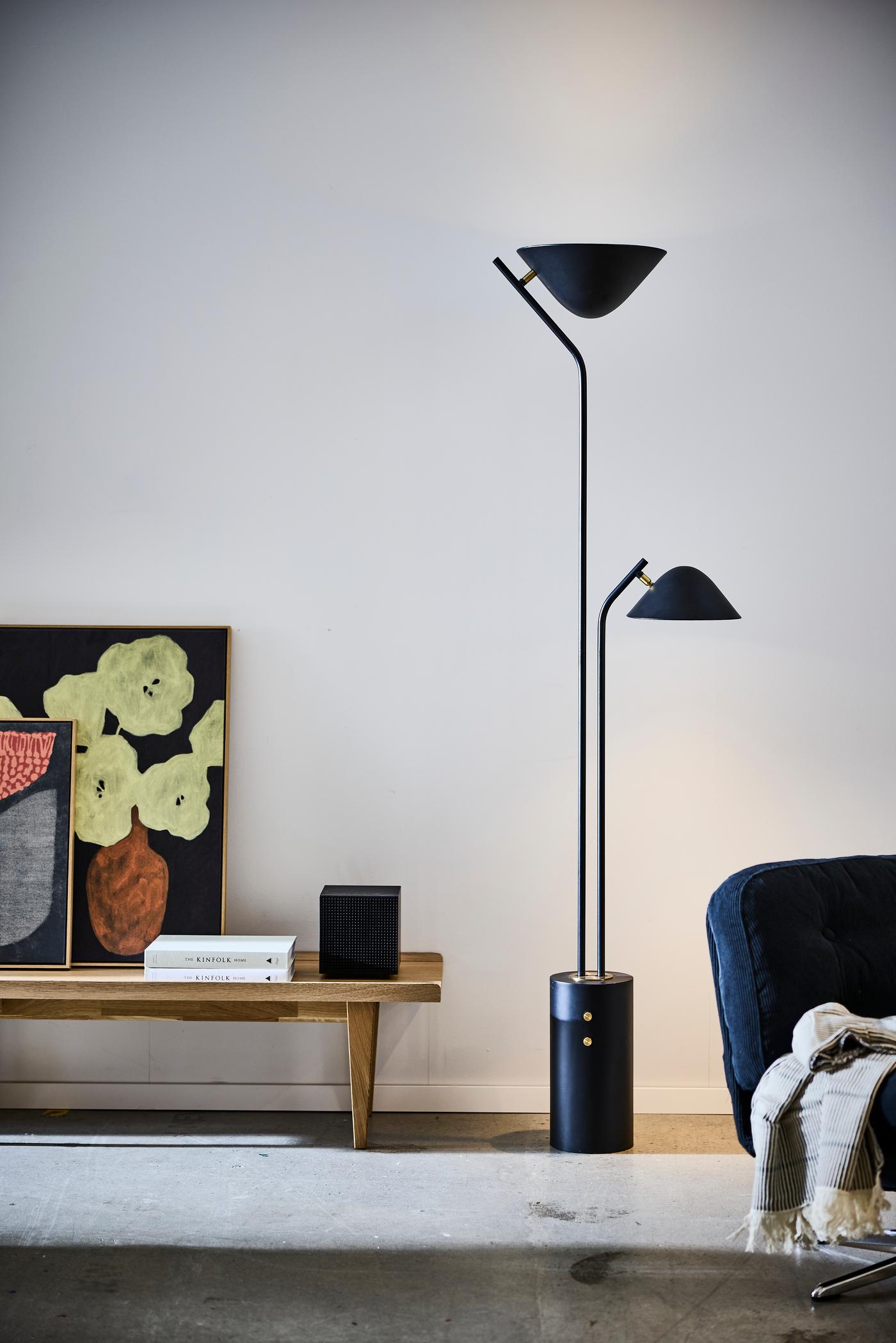 Black floor lamp with dual heads standing in a minimal living space beside a wooden bench, artwork, and chair.