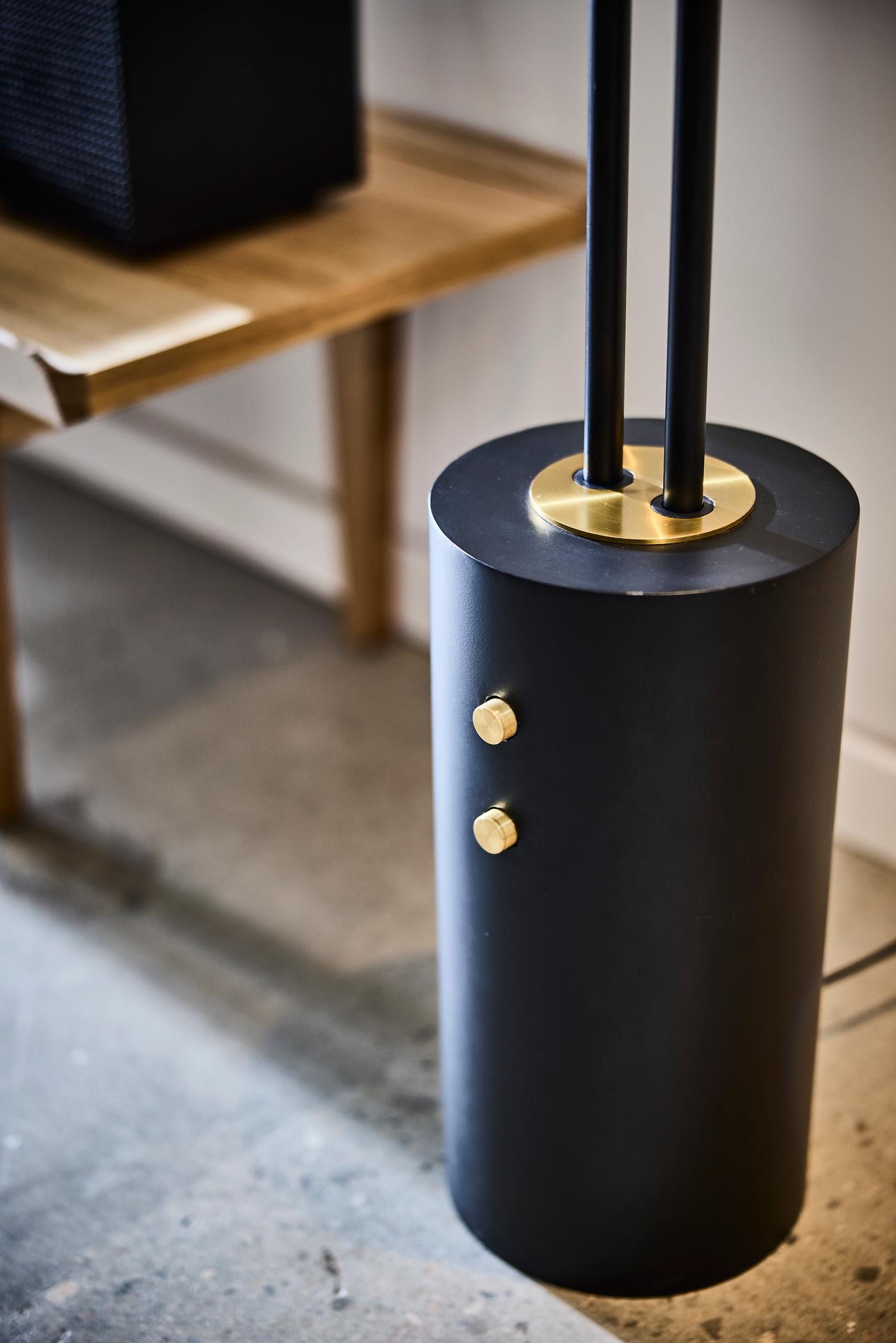Close-up of black floor lamp base with brass details and twin rods, with a wooden bench and speaker in the background.