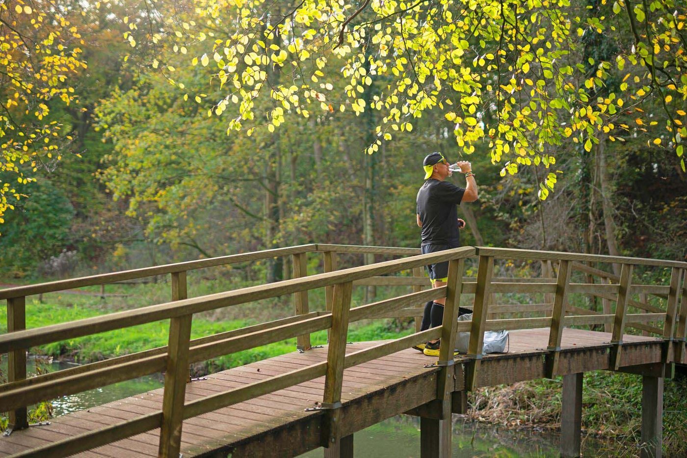 A man stands on a wooden bridge in a forest and drinks water.